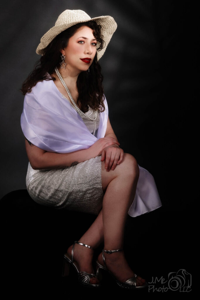 Alysia in a cream beaded dress, lilac wrap and wide-brimmed straw hat, seated against a dark ground