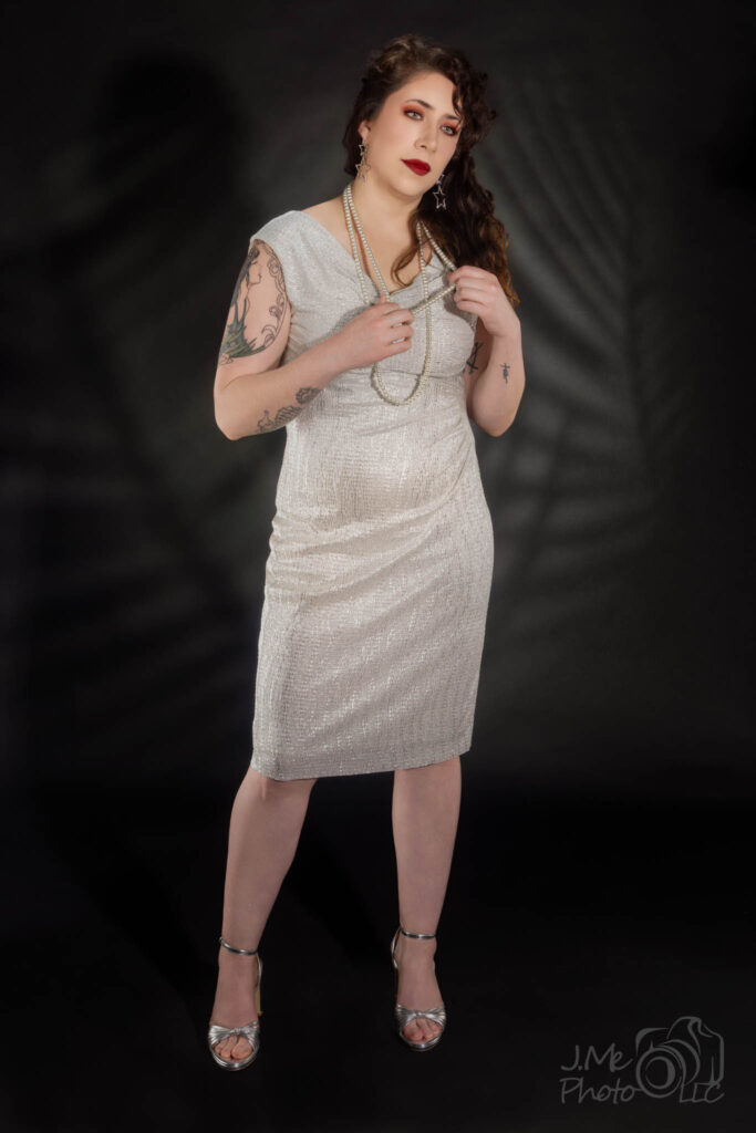 Alysia standing in a cream beaded dress with long pearls, a leaf-patterned shadow falling across the wall behind her