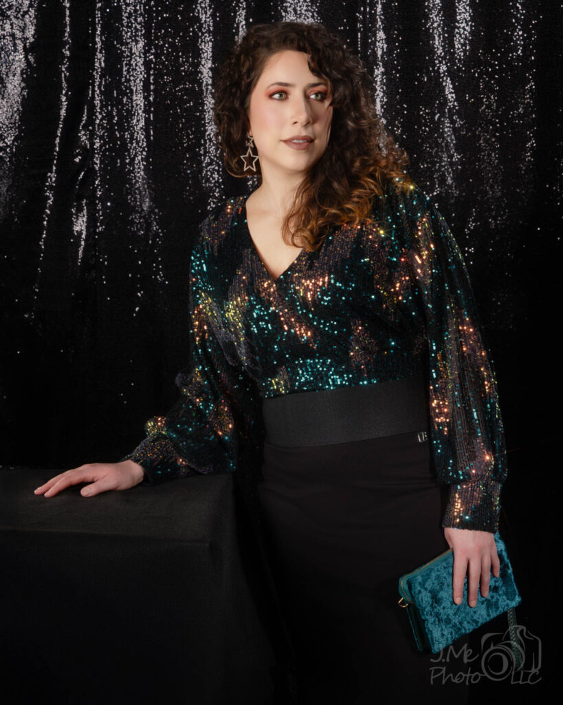 Alysia in an iridescent sequined blouse and black skirt, seated against a silver tinsel curtain