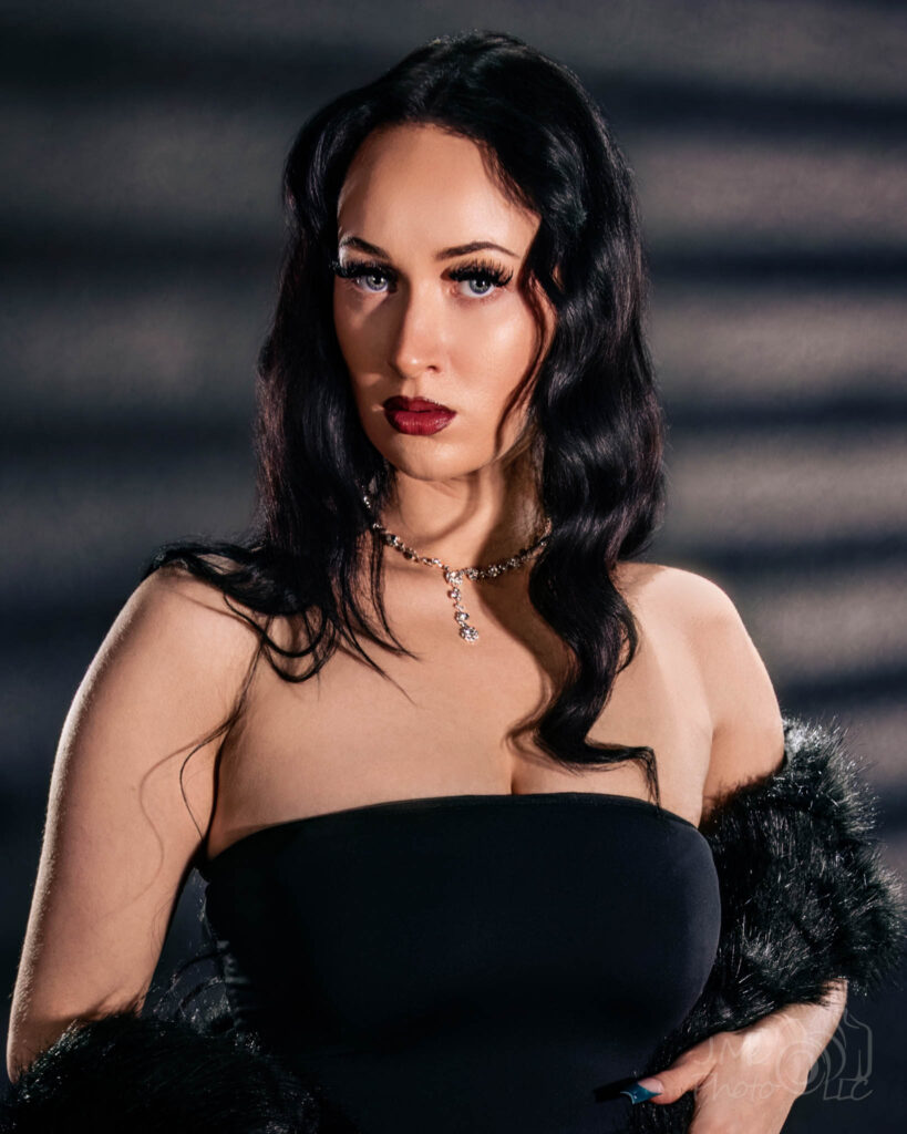 Autumn in a black strapless dress with black fur, hard slatted shadows across the wall behind her