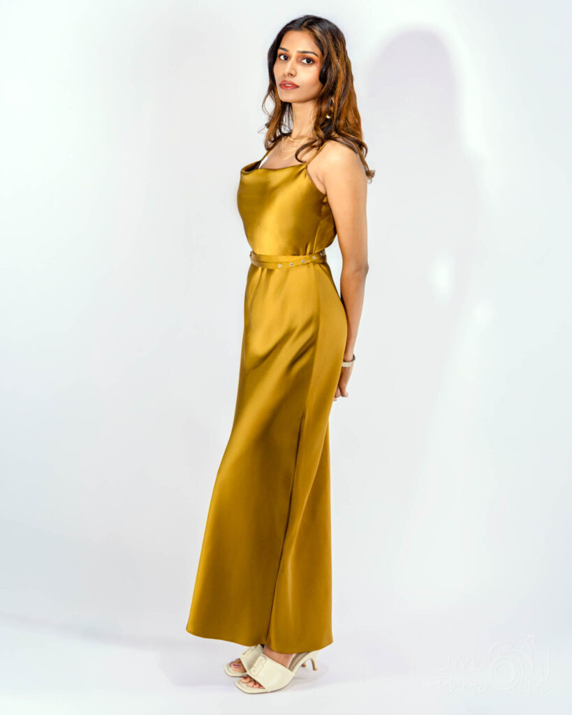 Srija standing in a gold satin gown against a plain pale wall, one arm at her side